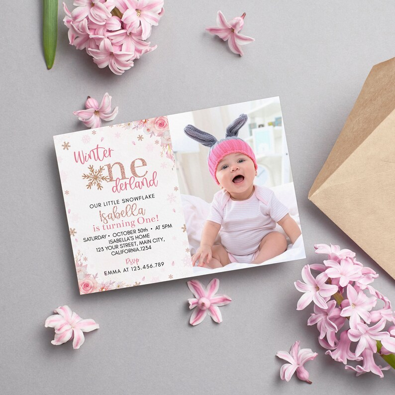 May include: A rectangular invitation card featuring a photo of a baby in a pink hat with bunny ears. The text reads Winter onederland, Our little snowflake, Isabella is turning One, Saturday October 30th at 5pm, Isabella's home, and RSVP Emma.