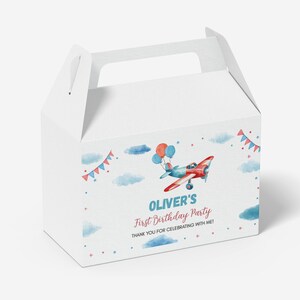 May include: A white cardboard gable box featuring a watercolor illustration of a red and blue airplane with balloons, clouds, and bunting. The text reads: OLIVER'S First Birthday Party THANK YOU FOR CELEBRATING WITH ME! The box has a top handle.