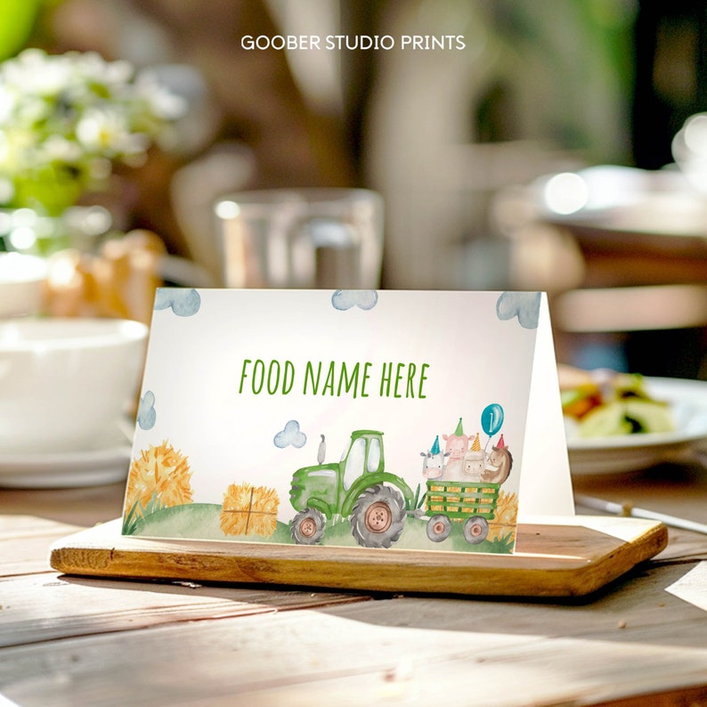May include: A printable food tent card featuring a green tractor with a hay bale and farm animals. The text "FOOD NAME HERE" is printed on the card.