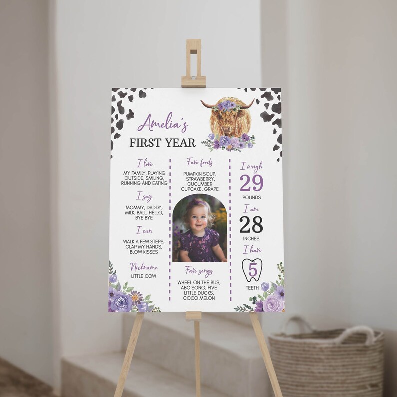 May include: A white milestone birthday sign for Amelia's first year featuring a highland cow illustration with purple flowers, black cow print borders, and text detailing favorite foods, songs, weight of 29 pounds, height of 28 inches, and 5 teeth.