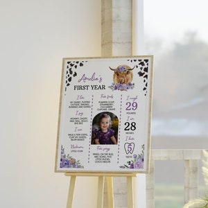 May include: A rectangular milestone sign featuring a highland cow illustration with purple flowers, black and white cow print borders, and text detailing Amelia's first year, including her weight of 29 pounds and height of 28 inches.