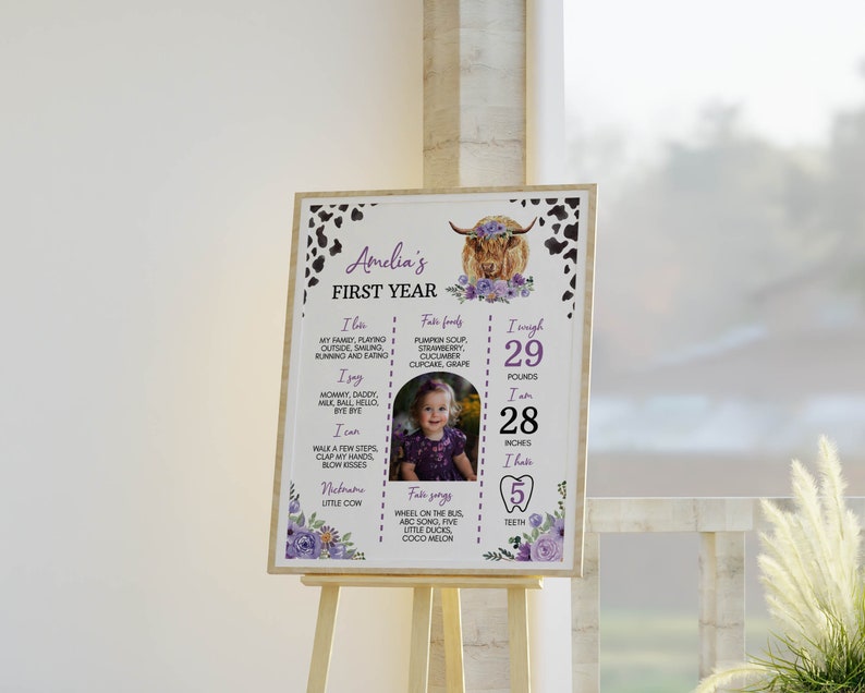 May include: A rectangular milestone sign featuring a highland cow illustration with purple flowers, black and white cow print borders, and text detailing Amelia's first year, including her weight of 29 pounds and height of 28 inches.