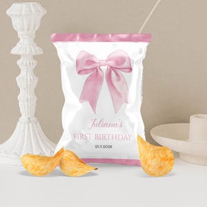 May include: A white snack bag with pink trim featuring a large pink bow graphic. The text reads Juliana's FIRST BIRTHDAY and 25.11.202X. Three golden potato chips are positioned in front of the bag, which is flanked by a white candle holder.