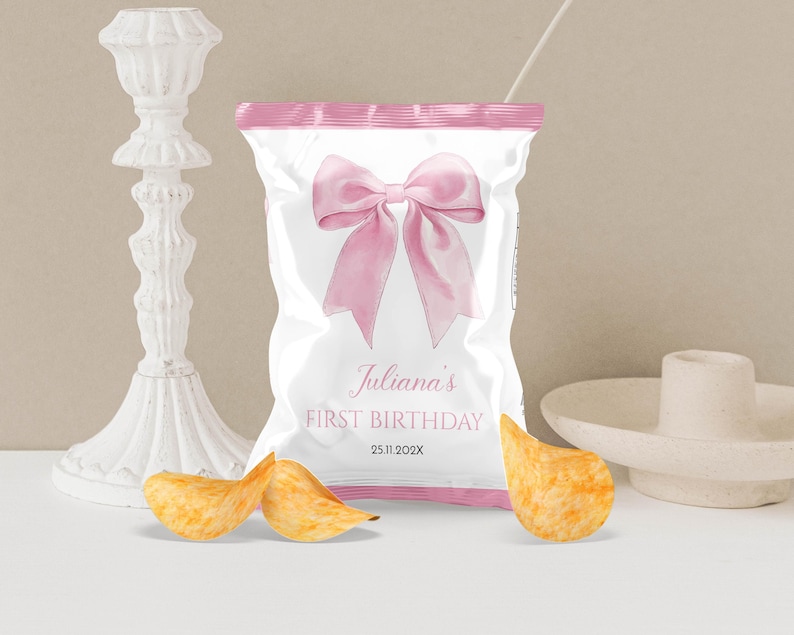 May include: A white snack bag with pink trim featuring a large pink bow graphic. The text reads Juliana's FIRST BIRTHDAY and 25.11.202X. Three golden potato chips are positioned in front of the bag, which is flanked by a white candle holder.
