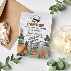 May include: A baby shower invitation with a woodland theme. The invitation features a brown bear, a tent, a campfire, and a squirrel. The text on the invitation reads "Here comes a little camper" and "Join us for a baby shower honoring Amelia Johnson".