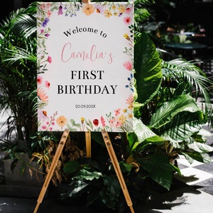 K&ouml;nnte beinhalten: Ein rechteckiges wei&szlig;es Schild auf einer Holzstaffelei mit einem Aquarell-Blumenrand in Rosa, Gelb, Lila und Gr&uuml;n. Der Text lautet: Welcome to Camelia's FIRST BIRTHDAY 20.09.202X. Das Schild ist vor einem Hintergrund aus gr&uuml;nem Blattwerk platziert.