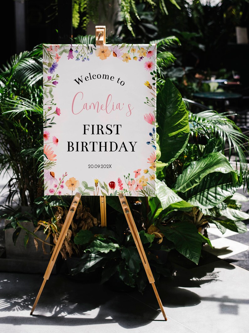 K&ouml;nnte beinhalten: Ein rechteckiges wei&szlig;es Schild auf einer Holzstaffelei mit einem Aquarell-Blumenrand in Rosa, Gelb, Lila und Gr&uuml;n. Der Text lautet: Welcome to Camelia's FIRST BIRTHDAY 20.09.202X. Das Schild ist vor einem Hintergrund aus gr&uuml;nem Blattwerk platziert.