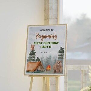 Op de afbeelding: Een rechthoekig bord op een houten ezel met de tekst WELCOME TO Benjamin's FIRST BIRTHDAY PARTY! 20.10.202X. Het ontwerp bevat aquarelillustraties van een bruine beer, een eekhoorn in een dennenboom, een tent en een kampvuur.