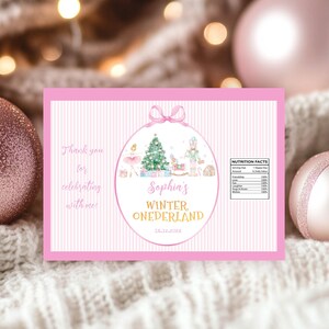 May include: A rectangular pink and white striped label featuring a central oval with a ballerina, Christmas tree, and nutcracker. Text reads: Thank you for celebrating with me!, Sophia's WINTER ONEDERLAND, 12.12.202X, and a Nutrition Facts table.