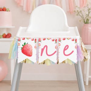 Puede incluir: Una pancarta blanca para trona con tres banderines, una ilustraci&oacute;n de fresa y las letras cursivas rosas O, N y E. La pancarta est&aacute; decorada con borlas de papel de seda en tonos pastel rosa, amarillo y azul.