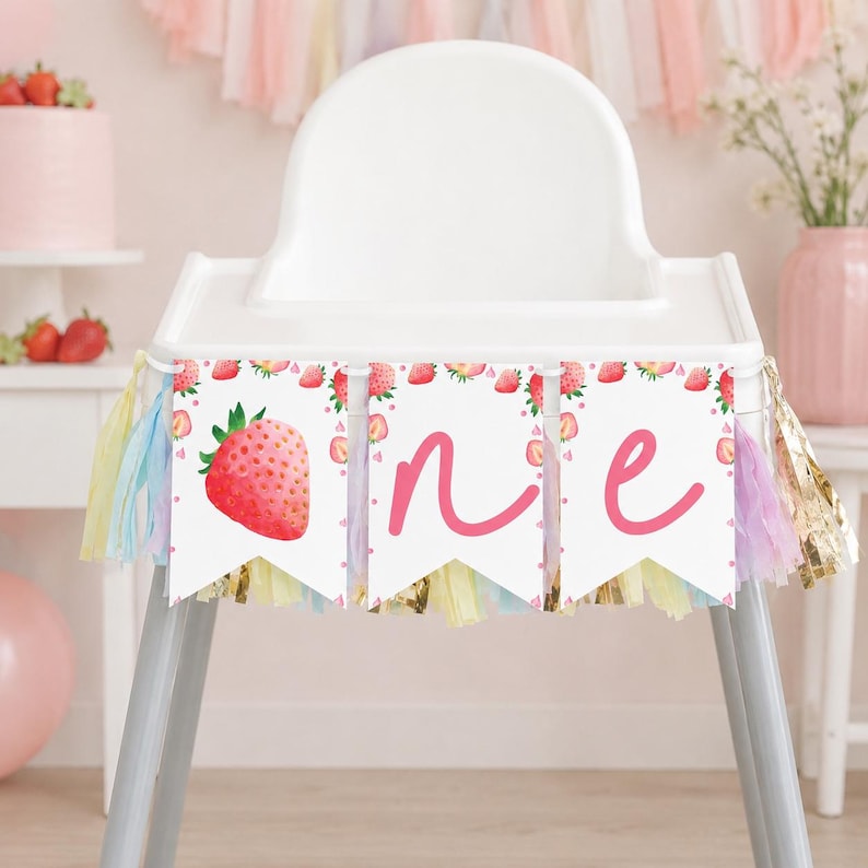 Puede incluir: Una pancarta blanca para trona con tres banderines, una ilustraci&oacute;n de fresa y las letras cursivas rosas O, N y E. La pancarta est&aacute; decorada con borlas de papel de seda en tonos pastel rosa, amarillo y azul.