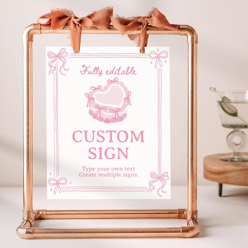 Peut inclure: Une pancarte rectangulaire avec des dessins au trait roses repr&eacute;sentant un g&acirc;teau en forme de c&oelig;ur et des n&oelig;uds, suspendue &agrave; un cadre en m&eacute;tal cuivr&eacute; par deux rubans en tissu marron. Le texte Fully editable, CUSTOM SIGN et Type your own text.