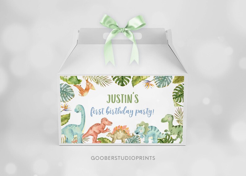 May include: A white gable box with a light green satin ribbon on top. The front features a watercolor illustration of various dinosaurs and tropical leaves surrounding the text "JUSTIN'S first birthday party!" in green and blue script.