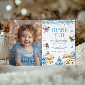 May include: A rectangular thank you card featuring a photo of a child in a blue dress on the left and text on the right. The text reads: THANK YOU, Thank you so much for celebrating my birthday with me and for the wonderful present you gave me! I had such a good time playing with all my friends and eating cake! Love, Amanda. The design includes blue fairy illustrations, snowflakes, and mushroom houses.