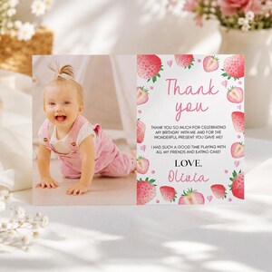 May include: A rectangular thank you card featuring a photograph of a baby in pink overalls on the left and a white background with pink strawberry illustrations and text on the right. The text reads Thank you, Olivia, and a message of gratitude.