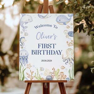 May include: A rectangular white birthday sign featuring watercolour illustrations of sea creatures including a dolphin, whale, turtle, octopus, jellyfish, and seahorse. The text reads Welcome To Oliver's FIRST BIRTHDAY and the date 20.06.202X.