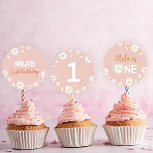 May include: Three cupcakes with pink frosting in white paper liners, each topped with a circular card featuring a daisy pattern on a light pink background. The cards display the text "MILA'S first birthday", the number "1", and "Mila is ONE".