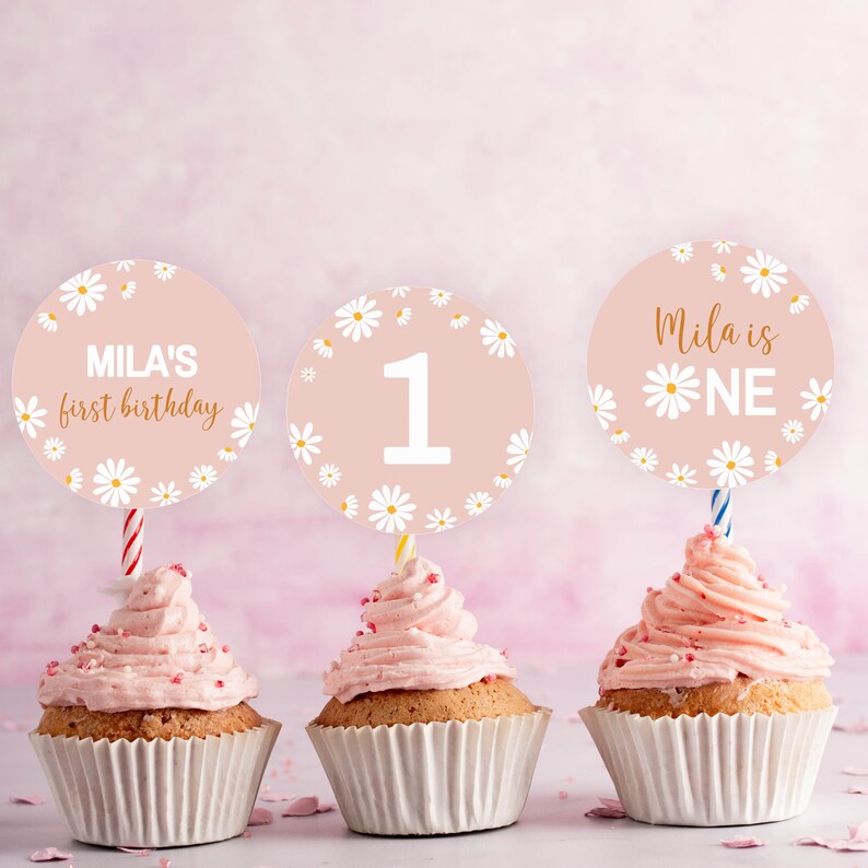 May include: Three cupcakes with pink frosting in white paper liners, each topped with a circular card featuring a daisy pattern on a light pink background. The cards display the text "MILA'S first birthday", the number "1", and "Mila is ONE".