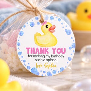 Pu&ograve; includere: Un'etichetta regalo rotonda bianca con una papera di gomma gialla con un fiocco rosa, bolle d'acqua blu e il testo THANK YOU for making my birthday such a splash! Love, Sophia. L'etichetta &egrave; attaccata a un sacchetto di plastica trasparente.
