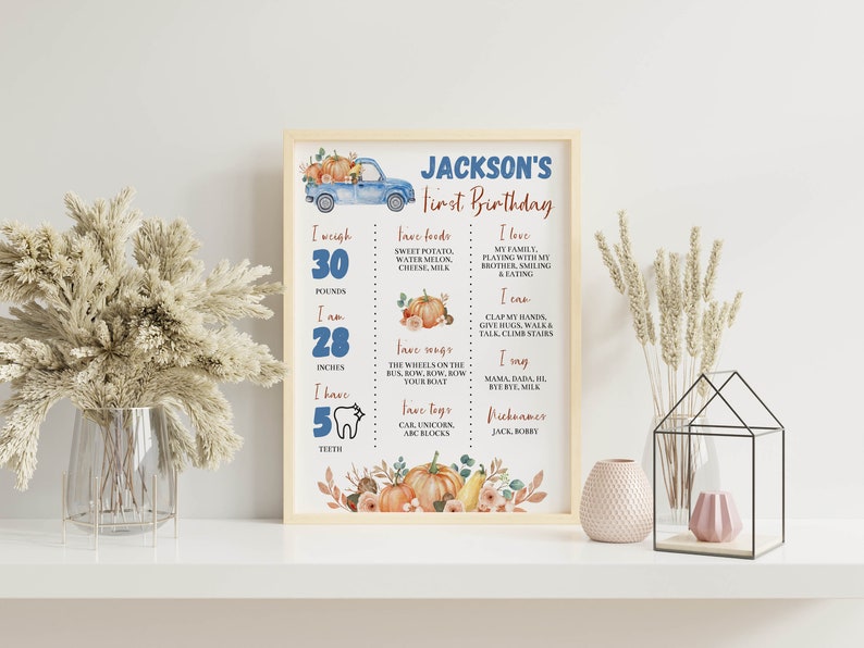 May include: A framed milestone poster for Jackson's first birthday featuring a blue truck, pumpkins, and text detailing weight of 30 pounds, height of 28 inches, 5 teeth, favorite foods, songs, toys, and personal milestones in blue and orange.