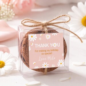 May include: A clear square plastic box containing a round chocolate cookie, tied with a brown twine bow. A square pink label features white daisy illustrations and the text: THANK YOU, For making my birthday so special!, Love, Mila.
