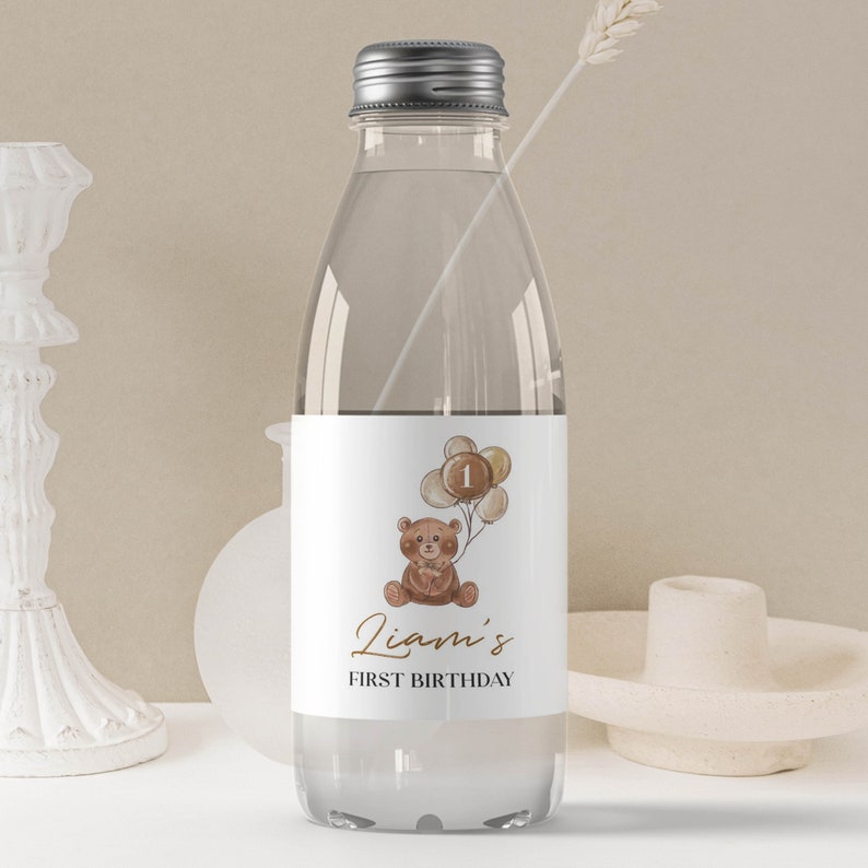 May include: A clear plastic water bottle with a white label featuring a brown teddy bear holding a cluster of balloons with the number one on the center balloon. The text on the label reads Liam's FIRST BIRTHDAY in brown and black lettering.