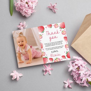 May include: A rectangular thank you card featuring a photo of a smiling toddler in pink overalls on the left and a white background with red strawberry illustrations and pink hearts on the right. The text reads Thank you, Love, Olivia.
