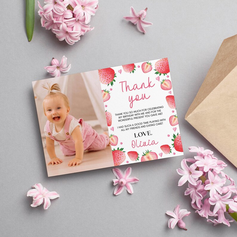 May include: A rectangular thank you card featuring a photo of a smiling toddler in pink overalls on the left and a white background with red strawberry illustrations and pink hearts on the right. The text reads Thank you, Love, Olivia.