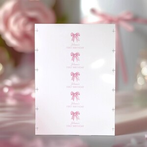 Puede incluir: Una hoja rectangular blanca con cinco repeticiones verticales de una ilustraci&oacute;n de un lazo rosa. Debajo de cada lazo, el texto dice Julianna's FIRST BIRTHDAY en una fuente cursiva rosa claro. Marcas de registro negras visibles en los bordes.