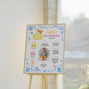 May include: A rectangular birthday milestone sign featuring a yellow rubber duck illustration, blue watercolor bubbles, and text including Sophia's 1st birthday, 20.08.202X, 30 pounds, 28 inches, 5 teeth, and a central photograph of a baby in a yellow outfit.