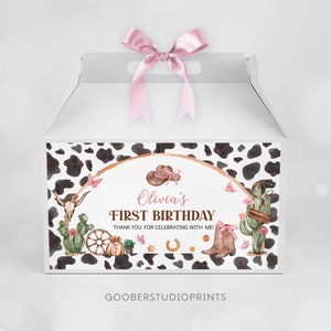 May include: A white cardboard favor box with a pink satin bow on top, featuring a cow print border and a central graphic with a cowboy hat, cactus, skull, and boots. The text reads: Olivia's FIRST BIRTHDAY THANK YOU FOR CELEBRATING WITH ME!