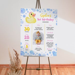 May include: A rectangular birthday milestone sign featuring a yellow rubber duck, blue watercolor bubbles, and pink text. It displays Sophia's 1st birthday details, including weight in pounds, height in inches, teeth count, and personal milestones.