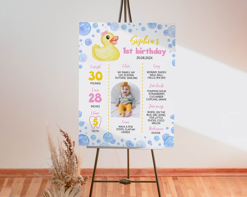 May include: A rectangular birthday milestone sign featuring a yellow rubber duck, blue watercolor bubbles, and pink text. It displays Sophia's 1st birthday details, including weight in pounds, height in inches, teeth count, and personal milestones.