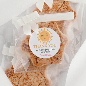 May include: Star-shaped cookies in clear plastic bags tied with white ribbon. Each bag features a round sticker with a yellow sun illustration and the text: THANK YOU for making my party so bright! Love, Olivia. The cookies have a textured brown surface.
