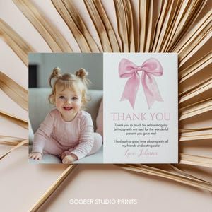 May include: A personalized thank you card featuring a photo of a smiling toddler in pink clothing. The card includes a watercolor illustration of a pink bow and the text "THANK YOU" with a handwritten message.
