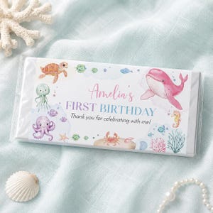 May include: A rectangular candy bar wrapper featuring a white background with watercolor illustrations of a pink whale, sea turtle, octopus, crab, and seahorse. The text reads Amelia's FIRST BIRTHDAY Thank you for celebrating with me!