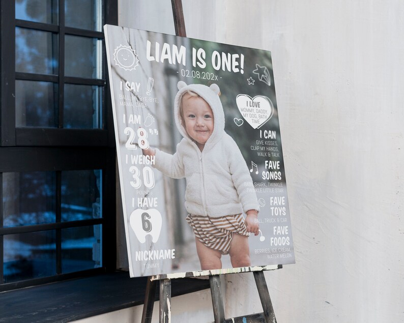 May include: A rectangular milestone birthday sign featuring a central photograph of a toddler in a white hooded jacket and striped shorts. The sign includes white text detailing Liam is one, birth date, weight of 30 pounds, 6 teeth, and favorites.