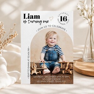 May include: A rectangular birthday invitation card featuring a photo of a child in denim overalls and a striped shirt sitting with wooden toy airplanes. The text reads Liam is turning one, Saturday December 16, 202X, Join us to celebrate, RSVP to Emma at 123.456.789, Saturday December 16th at 2pm, William's house, 123 Main Street, Texas. 1235.