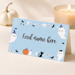 May include: A light blue folded card with the text Food name here in black script. The border features illustrations of white ghosts with purple hats, orange pumpkins, a black cat, spider webs, bats, skulls, stars, and candy pieces.