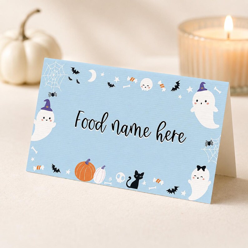 May include: A light blue folded card with the text Food name here in black script. The border features illustrations of white ghosts with purple hats, orange pumpkins, a black cat, spider webs, bats, skulls, stars, and candy pieces.