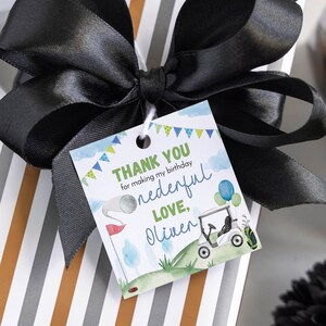 May include: A square gift tag with a golf theme featuring a golf cart, a golf ball on a tee, and blue and green bunting. The text reads: THANK YOU for making my birthday onederful LOVE, Oliver. The tag is tied with a large black satin ribbon.