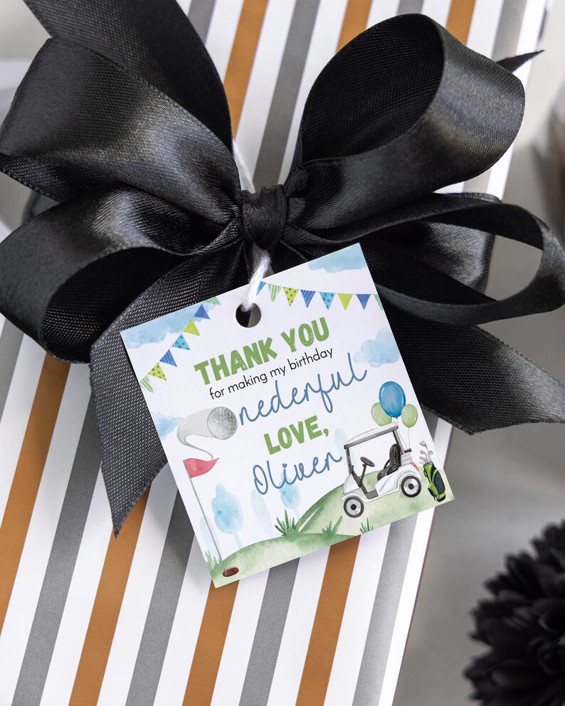 May include: A square gift tag with a golf theme featuring a golf cart, a golf ball on a tee, and blue and green bunting. The text reads: THANK YOU for making my birthday onederful LOVE, Oliver. The tag is tied with a large black satin ribbon.