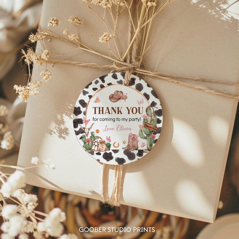 K&ouml;nnte beinhalten: Ein rundes Geschenkanh&auml;nger-Etikett mit einem schwarz-wei&szlig;en Kuhflecken-Rand. Die Mitte zeigt Illustrationen von einem Cowboyhut, Kaktus, Stiefeln und Schmetterlingen. Der Text lautet: THANK YOU for coming to my party! Love, Olivia. Mit Bindfaden.