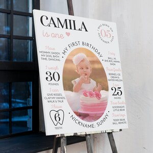 May include: A white rectangular birthday milestone sign featuring the name CAMILA, a central oval photo of a child with a cake, and text detailing birth date, weight of 30 pounds, height of 25 inches, 6 teeth, and favorite things.