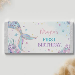 Op de afbeelding: Een rechthoekige wikkel voor een chocoladereep met een witte achtergrond en een holografische zeemeermin staart aan de linkerkant. De tekst luidt Mayra's FIRST BIRTHDAY in roze, turquoise en paars. Het ontwerp bevat aquarel zeedieren en sterren.