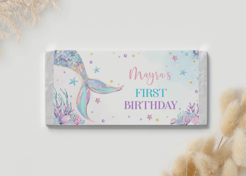 Op de afbeelding: Een rechthoekige wikkel voor een chocoladereep met een witte achtergrond en een holografische zeemeermin staart aan de linkerkant. De tekst luidt Mayra's FIRST BIRTHDAY in roze, turquoise en paars. Het ontwerp bevat aquarel zeedieren en sterren.