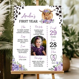 May include: A rectangular milestone sign featuring a cow print border, a highland cow illustration with purple flowers, and text detailing Amelia's first year, including her weight of 29 pounds, height of 28 inches, and 5 teeth, with a central photo.