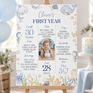 May include: A white milestone birthday sign titled Oliver's FIRST YEAR featuring blue text, a central photo of a child, and watercolor illustrations of sea creatures including a dolphin, whale, octopus, jellyfish, and seahorses with ocean plants.