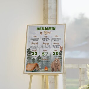 May include: A rectangular milestone birthday sign for a one-year-old named Benjamin, featuring a forest theme with a bear, tent, and campfire. The text lists milestones including weight in pounds, height in inches, favorite foods, and songs.