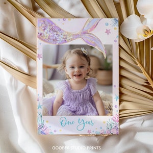 Pode incluir: Um adere&ccedil;o de cabine de fotos imprim&iacute;vel com uma cauda de sereia com glitter roxo e dourado, um fundo rosa e o texto "One Year".